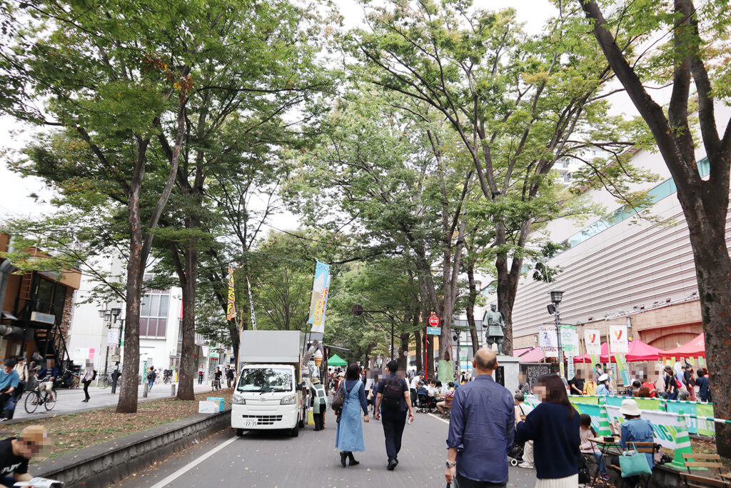 木々に囲まれた通りで行われているイベントの様子です。歩行者天国となった道路には、人々が散策したり、出店を楽しんでいます。白い車両が停まり、奥にはピンク色のテントが並び、賑やかな雰囲気が漂っています。通りの両脇にはイベントの旗が掲げられ、音楽祭やお祭りのような雰囲気を感じさせます。