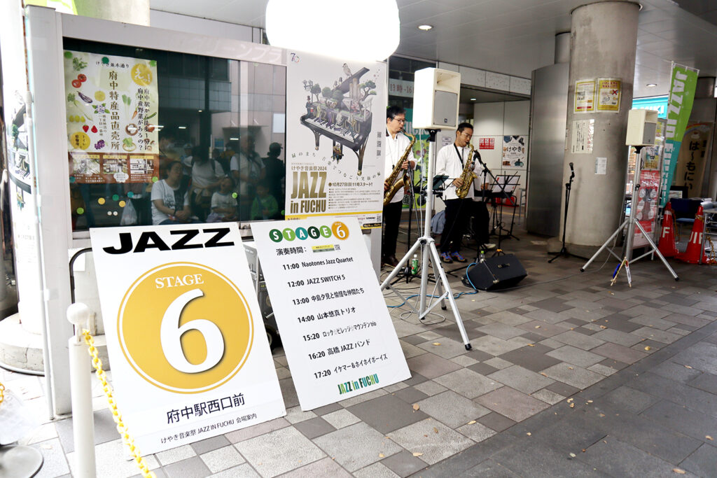 「JAZZ in FUCHU 2024」のイベント会場で、府中駅西口に設置された「STAGE 6」の看板が目立っています。サックス奏者たちがライブ演奏を行い、観客は楽しそうに聴き入っています。看板にはステージスケジュールが記載され、複数のジャズバンドが参加することが示されています。イベントの活気あふれる雰囲気が伝わる一枚です。