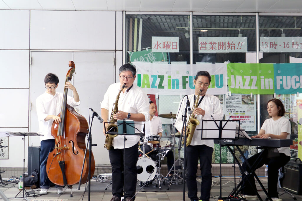 白いシャツを着た5人のジャズバンドが「JAZZ in FUCHU 2024」の会場で演奏を行っています。ベース、サックス、ドラム、キーボードといった楽器を使用し、観客に生演奏を披露しています。後ろの横断幕にはイベント名が描かれ、活気ある会場の雰囲気が伝わります。