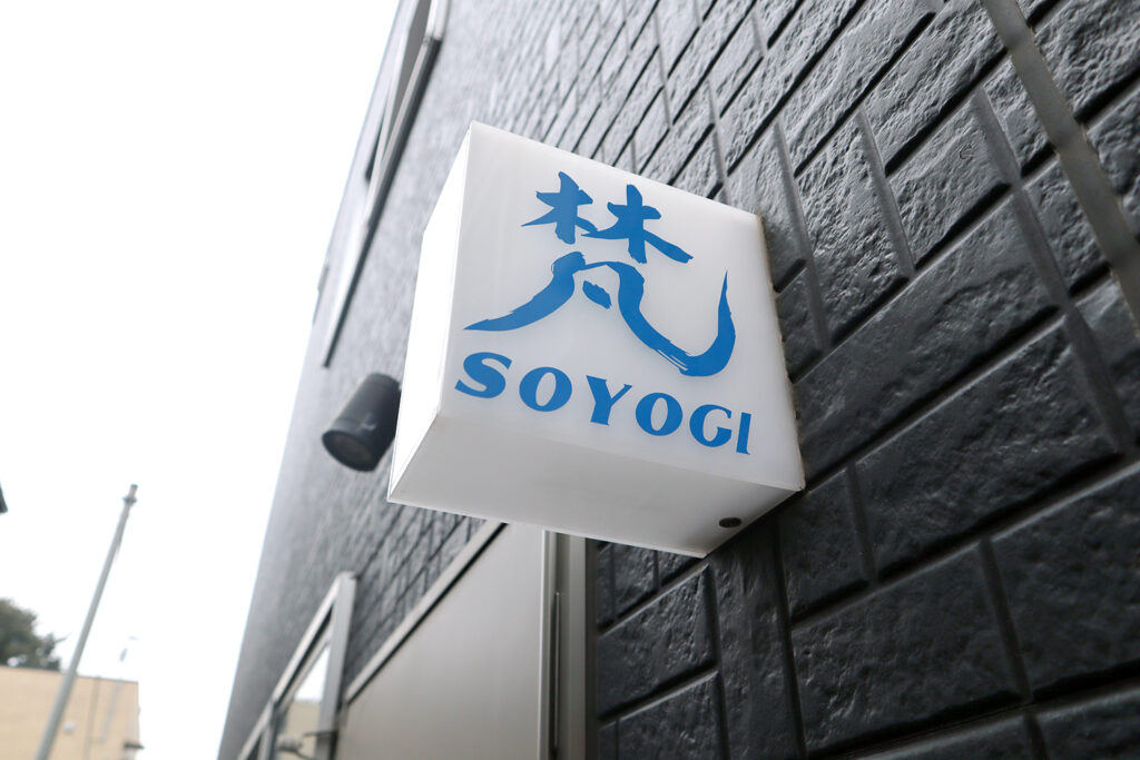 黒い壁に取り付けられた「SOYOGI」という店の看板の写真です。看板には青い筆書き風の漢字「楚」の文字が目立ち、その下に「SOYOGI」とローマ字で表記されています。