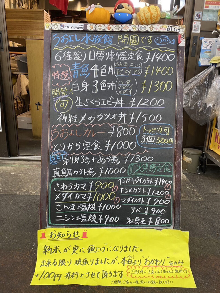 店舗前に置かれた「おさかな食堂 うおよし」の黒板メニュー。手書きで鮮魚定食や丼もの、焼き魚などの詳細が記載されており、価格がわかりやすく表示されている。黒板上部には季節感のある飾り付けがされ、黄色い紙には白米値上げの案内が書かれている。手作り感のある親しみやすい雰囲気が感じられるメニュー板。