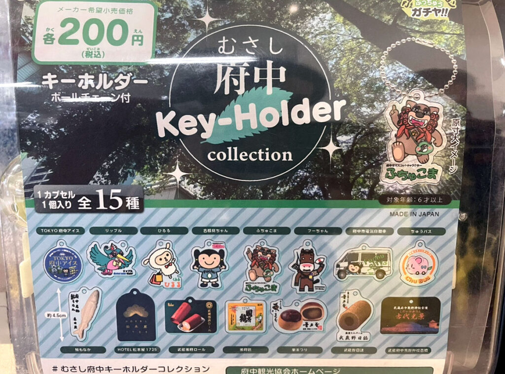 「むさし府中 Key-Holder collection」の商品の詳細が写された写真。15種類のデザインが紹介されており、「ふちゅこま」や「ちゅうバス」など府中市のキャラクターや名物をモチーフにしたキーホルダーが並んでいます。価格は税込200円で、1カプセルに1個が入っています。「MADE IN JAPAN」と記載されており、品質が強調されています。背景には木漏れ日が映え、自然を感じさせるデザインになっています。