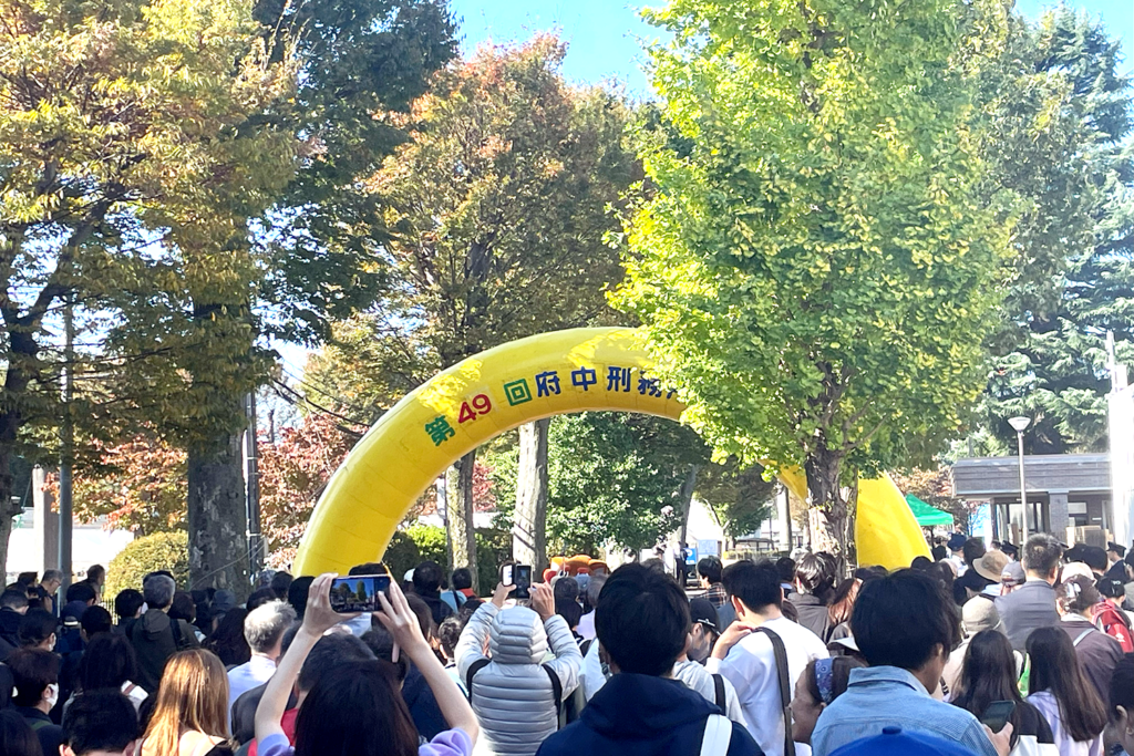 [「第49回府中刑務所文化祭」の入り口に設置された黄色いアーチ型のゲートです。ゲートには青と赤で「第49回府中刑務所文化祭」と書かれており、多くの来場者が並んでいる様子が見られます。背景には青空と緑の木々が広がり、晴れた日にイベントが開催されていることがわかります。