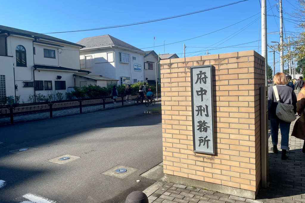 府中刑務所の入口近くにあるレンガ造りの看板で、「府中刑務所」と書かれた標識が設置されています。周囲には歩行者や自転車に乗った人々が通り、晴れた日の穏やかな街並みが広がっています。背景には住宅や電柱が見え、日常の風景の中に刑務所の存在が溶け込んでいます。