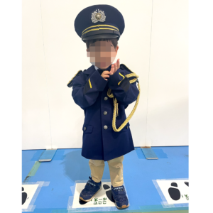 制服を着て敬礼のポーズをしている子供の写真です。子供は帽子と制服を身に着けており、肩には金色のロープ飾りが付いています。制服は少し大きめで、子供の体に少しぶかぶかの様子が微笑ましいです。背景は白い壁で、足元には立ち位置を示す足形のマークが貼られています。