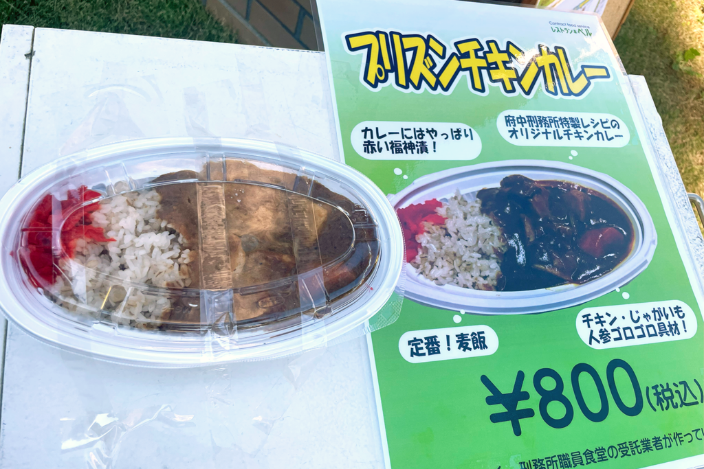 府中刑務所文化祭で販売されている「プリズンチキンカレー」の弁当写真です。容器には麦飯とチキンカレーが盛られ、福神漬けも添えられています。隣のポップには「府中刑務所特製レシピのオリジナルチキンカレー」と説明され、具材としてチキンやじゃがいも、にんじんが使われていると記載されています。価格は800円（税込）です。