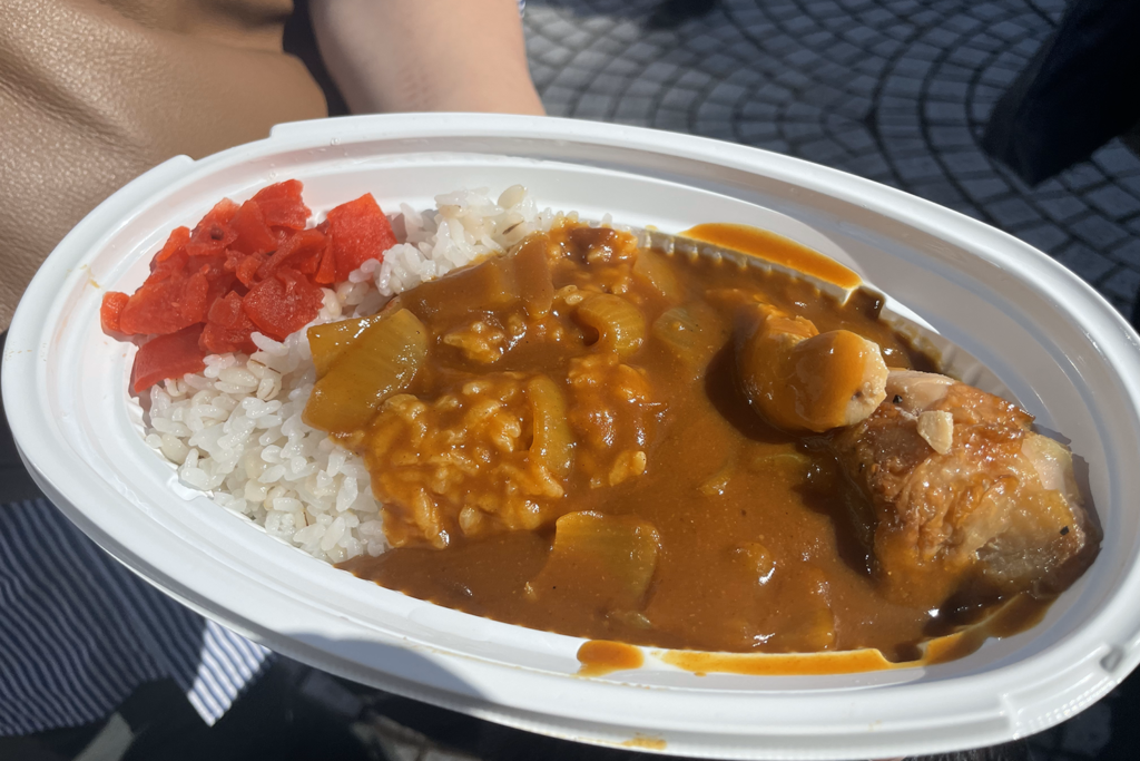 プラスチック容器に盛られたチキンカレーの写真です。麦ご飯の上にカレーがかけられ、左側には赤い福神漬けが添えられています。具材として大きめのチキンや野菜が入っており、ボリュームが感じられます。屋外で撮影されており、明るい日差しが料理を照らしています。