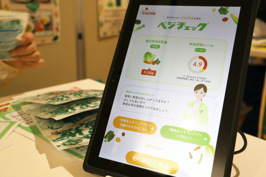 タブレットの画面に「ベジチェック」の測定結果が表示されています。画面には、推定野菜摂取量が「約250g」、野菜摂取レベルが「4.9」と示されています。グラフやイラストが用いられており、視覚的に結果が分かりやすくデザインされています。下部には、「野菜を摂る習慣をつけましょう」などのアドバイスが記載され、関連するアプリの案内ボタンも配置されています。背景には測定イベント用のチラシが数枚置かれています。