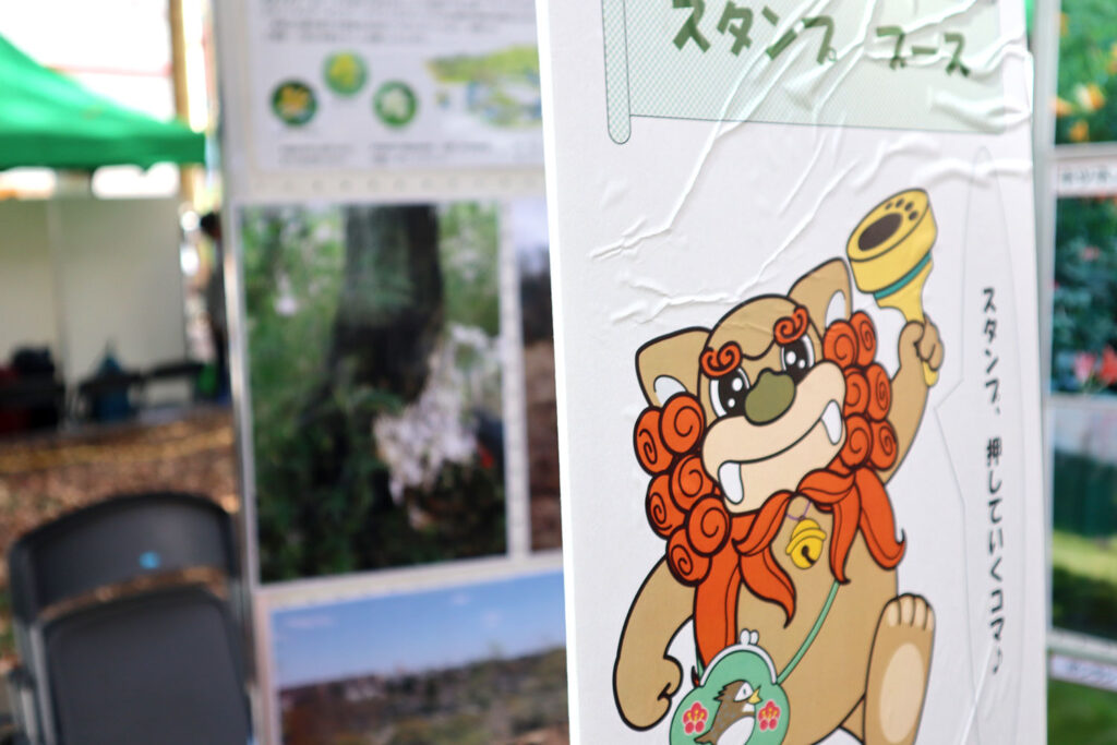 公園のイベントで設置されたスタンプコースの案内看板。赤いたてがみと可愛らしい表情のキャラクターが描かれており、手にはスタンプを持っています。背景には自然の写真パネルや緑のテントが見えます。