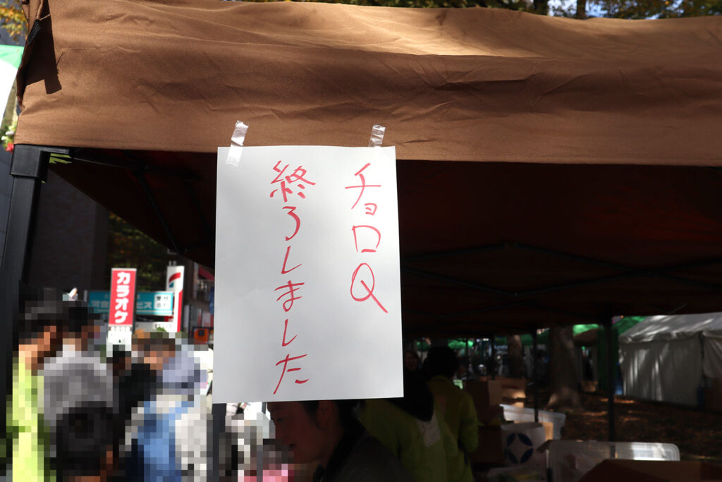 茶色のテントに貼られた「チョロQ終了しました」と手書きで書かれた白い紙。背景にはイベントのブースや人々が見えます。