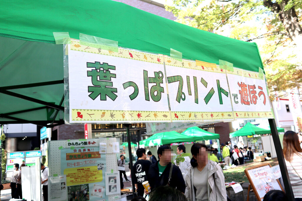 緑色のテントに取り付けられた「葉っぱプリントで遊ぼう」と書かれた看板が目立つ様子。看板は秋らしいデザインで、葉っぱや木のイラストが装飾されている。背景にはイベントの様子が広がり、参加者たちが楽しんでいる姿が見られる。