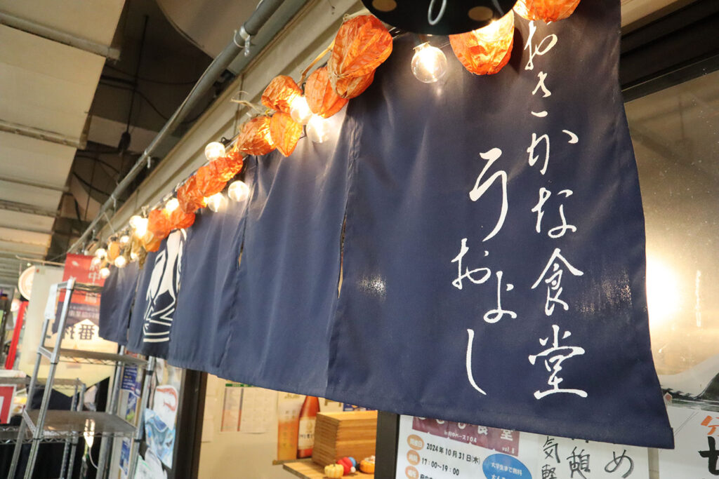 青い暖簾に「おさかな食堂 うおよし」と白い文字で書かれたデザイン。暖簾の上部には提灯風の飾りとライトが飾られ、温かみのある雰囲気が感じられる。背景には店内の一部やポスターが見える。