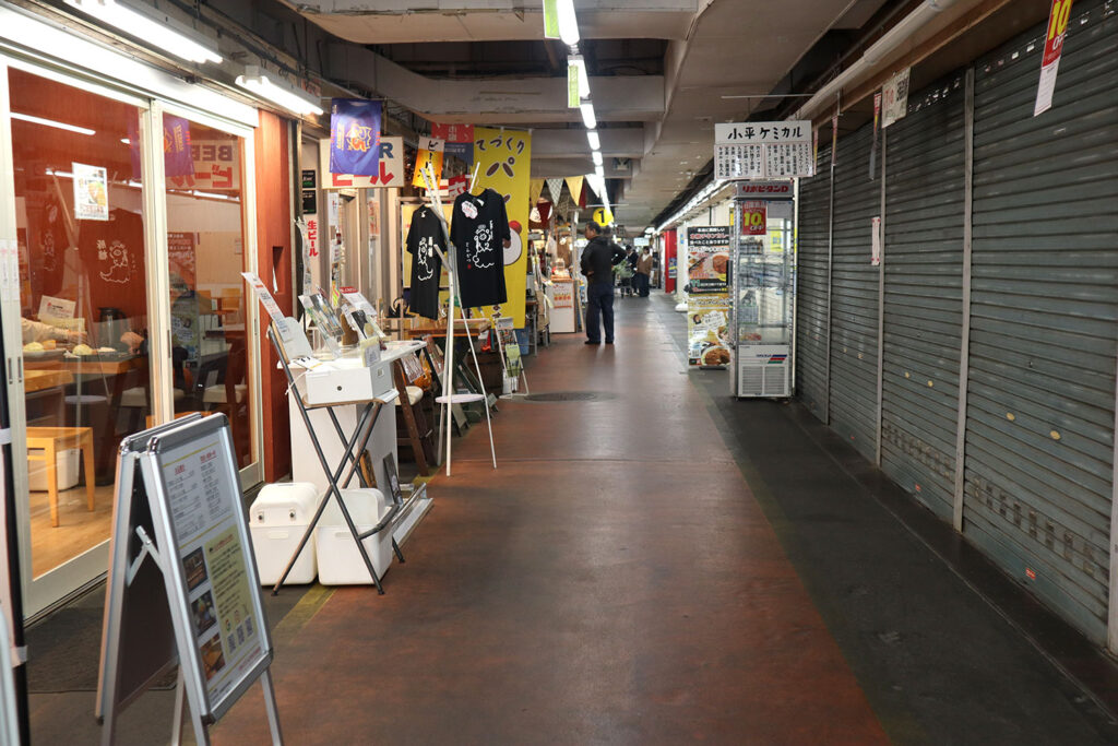 府中市場内の通路の様子。左側にはガラス張りの店舗や販売ブースが並び、手作り商品やポスターが飾られている。右側には閉店中のシャッターが目立つ。中央奥には歩く人の姿が見える。市場独特の雰囲気と活気を感じる通路。