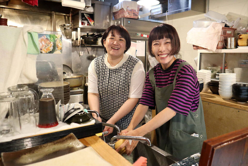 厨房で作業中の2人の女性スタッフが笑顔を見せている様子。手前には洗浄中の食器や調理器具が見える。背景には調理場の道具や棚に並んだ食器が並び、温かみのある雰囲気を感じさせる。