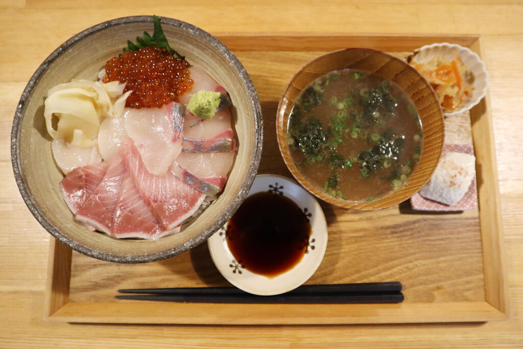 木製のトレーに盛られた定食の写真。新鮮な刺身が美しく盛り付けられた海鮮丼、海藻が入った味噌汁、小皿に盛られた副菜、醤油の小皿、そして箸がセットされている。シンプルながらも丁寧に準備された和食の一膳。