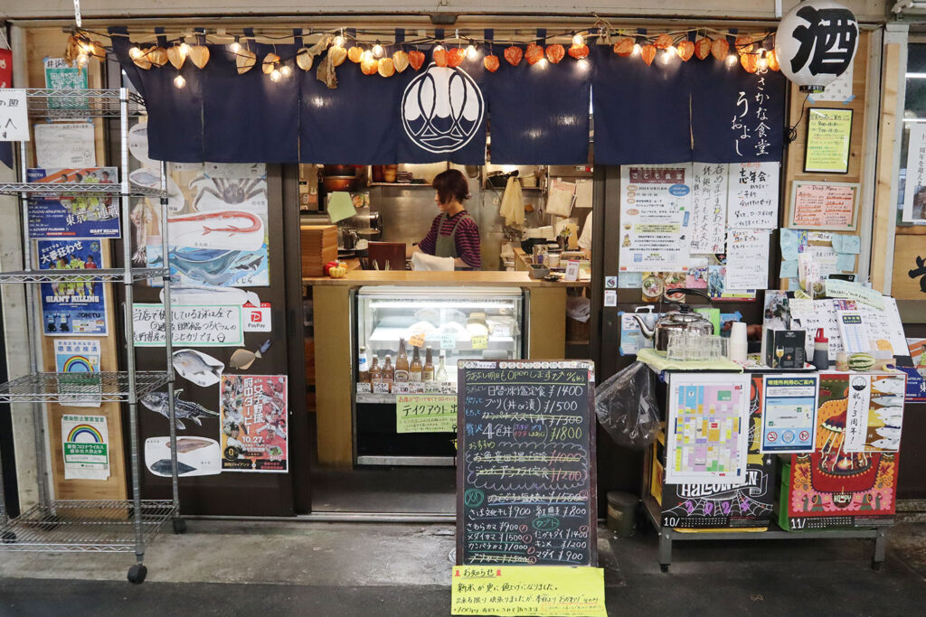 食堂の外観写真。青い暖簾に店名のロゴが描かれ、上部には提灯や飾りが吊るされている。店頭には黒板メニューが置かれ、旬の魚介料理の価格や内容が記載されている。ショーケースには鮮魚や商品が並び、店内ではスタッフが作業中。温かみのある雰囲気の店構え。