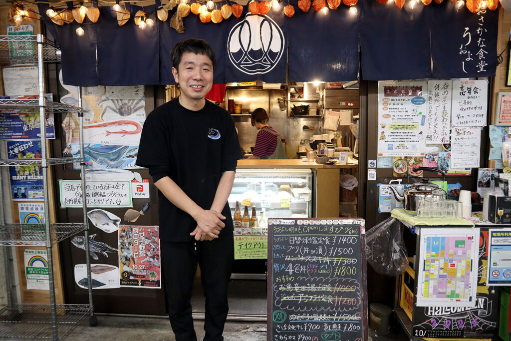 「おさかな食堂 うおよし」の店舗前で笑顔で立つ男性店員。店の暖簾には魚のデザインが描かれており、上部に提灯型の飾りが点灯している。入口には魚のポスターや黒板メニューが掲示されており、日替わり定食や丼ものの価格が書かれている。店内には調理中のスタッフの姿が見える、家庭的で温かい雰囲気の食堂。