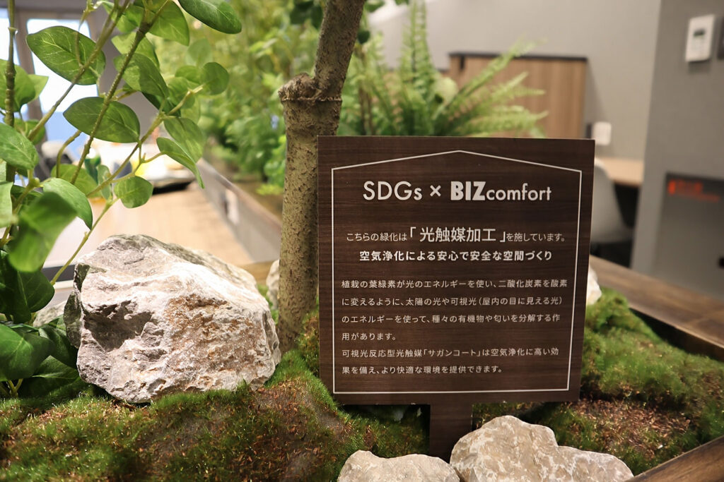 室内の緑化スペースに設置された「SDGs × BIZcomfort」と書かれた木製の案内板。板には「光触媒加工」による空気浄化の取り組みが記載されている。周囲にはリアルな植物と苔、白い石が配置され、自然を感じさせるデザインが施されている。背景にはオフィススペースがぼんやりと見える。