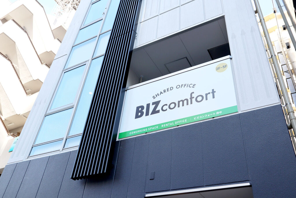 ビルの外壁に掲示された「BIZcomfort」の看板が目立つ写真。看板には「SHARED OFFICE」と「BIZcomfort」のロゴが表示され、その下には「COWORKING SPACE」「RENTAL OFFICE」「ビズコンフォート府中」と記載されている。背景にはビルの窓や外壁、モダンなデザインの建築要素が映り込んでおり、明るい日差しが降り注いでいる。