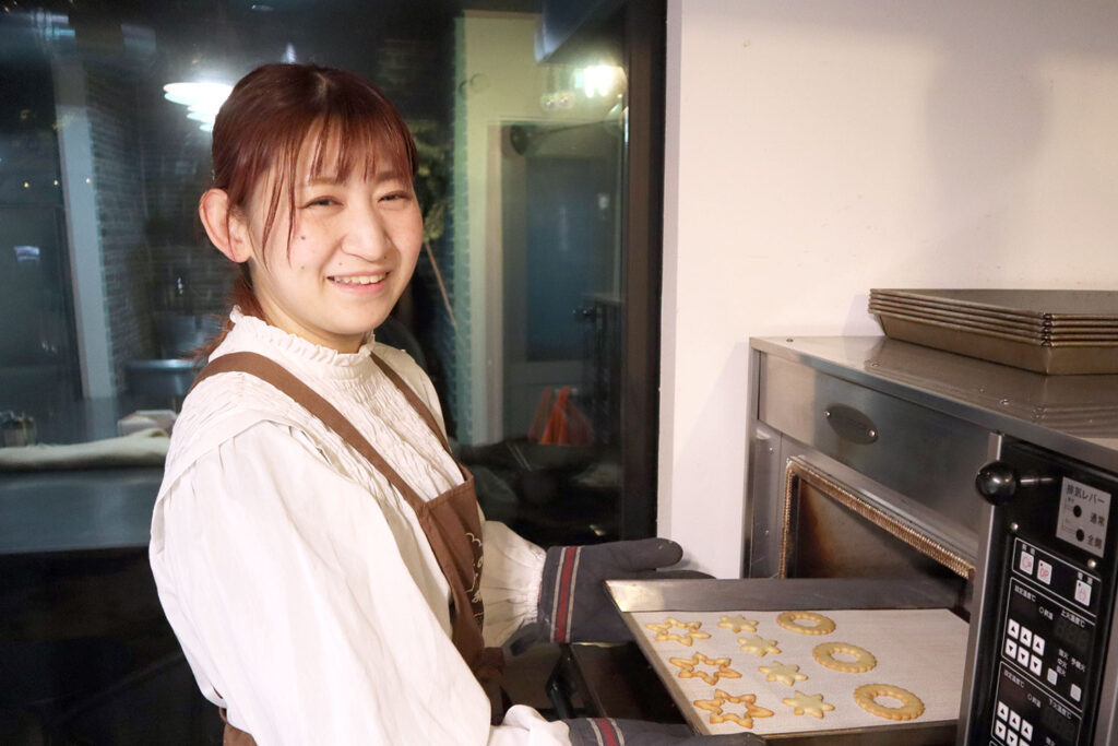 明るい笑顔でオーブンから焼きたてのクッキーを取り出している女性の写真。星型やリース型のクッキーが並ぶ天板を、厚手のミトンを着けた手で持っている。背景にはシンプルな厨房が映り、クッキー作りの温かい雰囲気が伝わってくる。白いブラウスと茶色のエプロンが清潔感と親しみやすさを演出している。