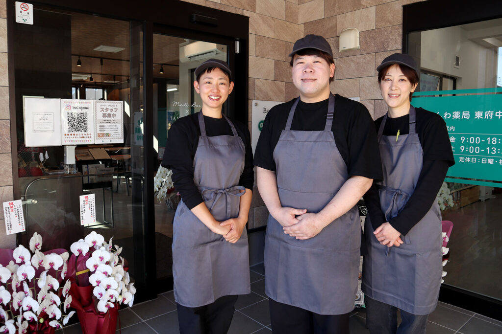 店舗の入口前で笑顔のスタッフ3人が並んでいる写真。全員が黒いシャツにグレーのエプロンとキャップを着用。背景には店舗のガラスドアがあり、店名や営業時間が書かれた緑色の看板が見える。足元には白い胡蝶蘭の鉢植えが並べられている。