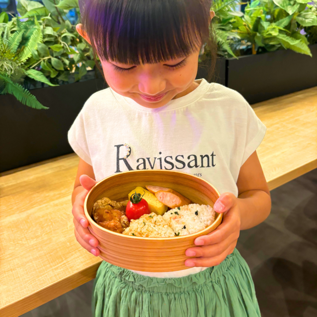 白い「Ravissant」とプリントされたTシャツを着た女の子が、木製の弁当箱を持ちながら微笑んでいる様子。弁当箱にはおにぎり、唐揚げ、ソーセージ、ミニトマトなどが詰められている。背景には木製のテーブルと緑の植物が並んでいる。