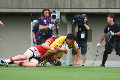 ラグビー試合中のタックルシーン。黄色いユニフォームを着た選手がボールを持ち、赤いユニフォームの選手にタックルされながら地面に倒れ込む瞬間が映されている。背景にはカメラマンや撮影機材を持つスタッフが試合の様子を撮影している姿が確認できる。激しい試合の一瞬を捉えた写真。