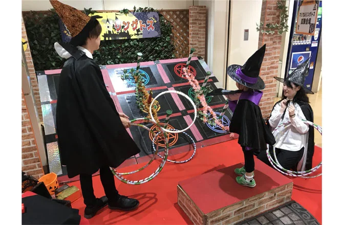 ハロウィンをテーマにした輪投げゲームの様子。魔女の帽子をかぶった子どもたちと大人が輪投げを楽しんでいます。背景にはカラフルな的と「トリックオアトス」と書かれた看板が飾られており、ハロウィンの雰囲気が演出されています。参加者全員が魔法使いの衣装を着てゲームに参加しています。