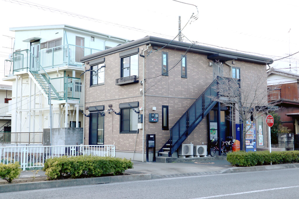 道路沿いにある2階建ての住宅兼店舗の建物。レンガ調の外壁に黒い階段が特徴的で、右下には自動販売機と看板が見えます。隣には淡い水色のスチール製階段を備えた別の建物があります。手前には植え込みが整備され、落ち着いた住宅街の一角が描かれています。