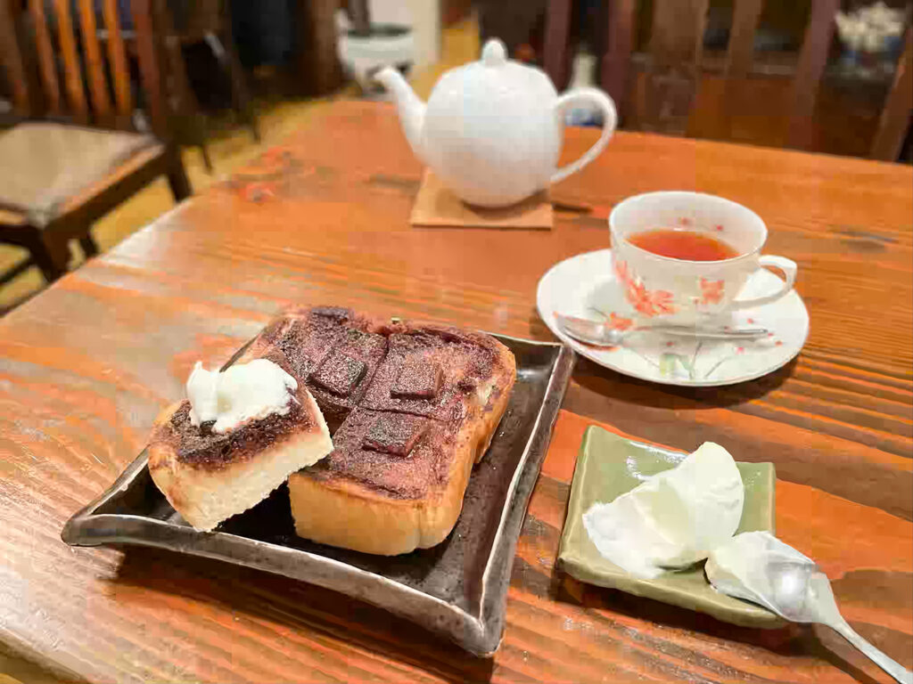 木製テーブルに置かれたトーストと紅茶のセット。トーストにはチョコレートがかかり、ホイップクリームが添えられています。紅茶は花柄のティーカップに注がれ、奥には白いティーポットが見えます。