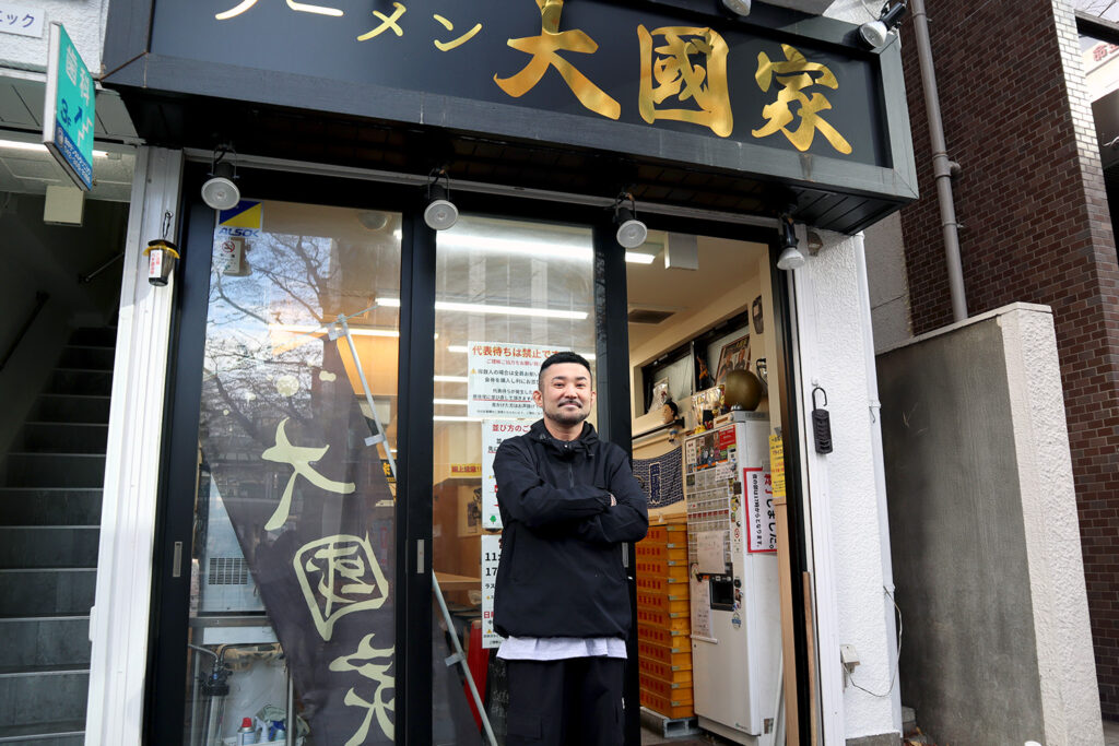 ラーメン店「大國家」の店頭で腕を組んで立つ男性の写真。黒いジャケットを着た男性が微笑みながら自信に満ちた様子でポーズを取っている。店舗の看板には金色の文字で「大國家」と書かれ、店内にはメニューや装飾が見える。右側には入り口の暖簾が垂れ下がり、左側には階段が写っている。温かみと親しみやすさを感じる店の外観が特徴的。