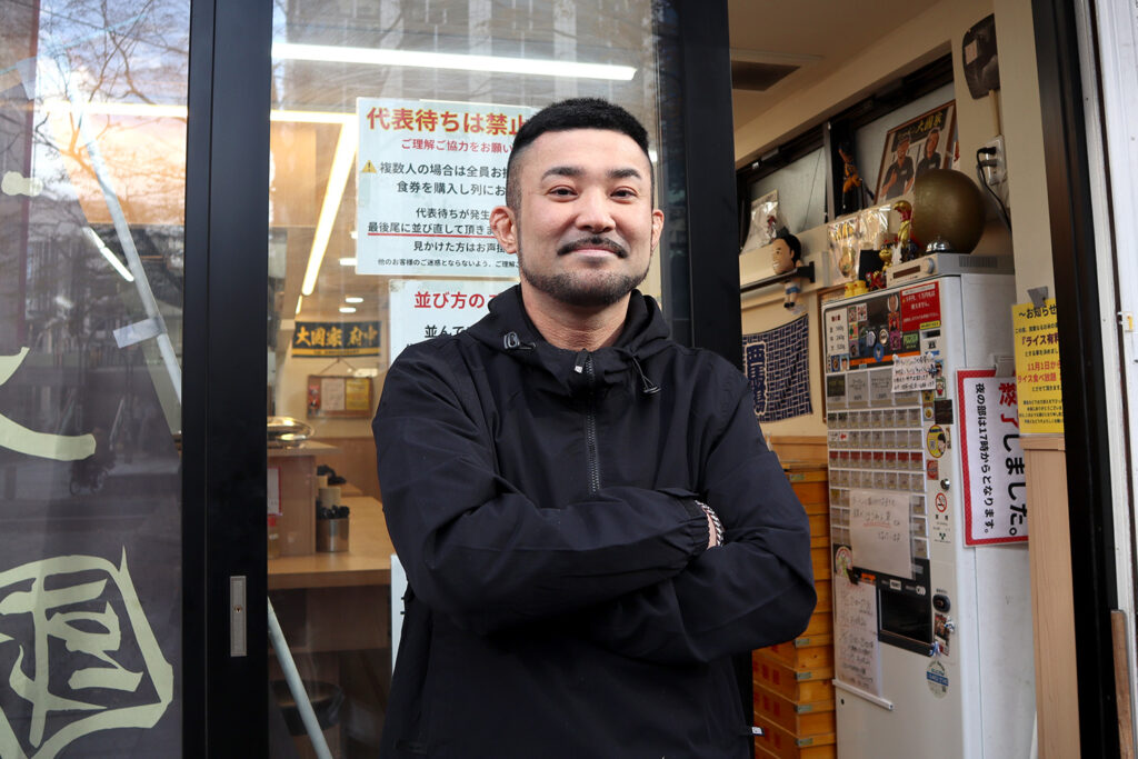 ラーメン店「大國家」の店頭で腕を組む男性のポートレート。黒いジャケットを着た男性が微笑み、自信に満ちた表情を見せている。背景には店内の様子が映り、食券機や装飾品、注意書きの掲示物が見える。ガラス扉には「大國家」の文字が反射し、温かみと親しみやすさを感じさせる店舗の雰囲気が伝わる写真。