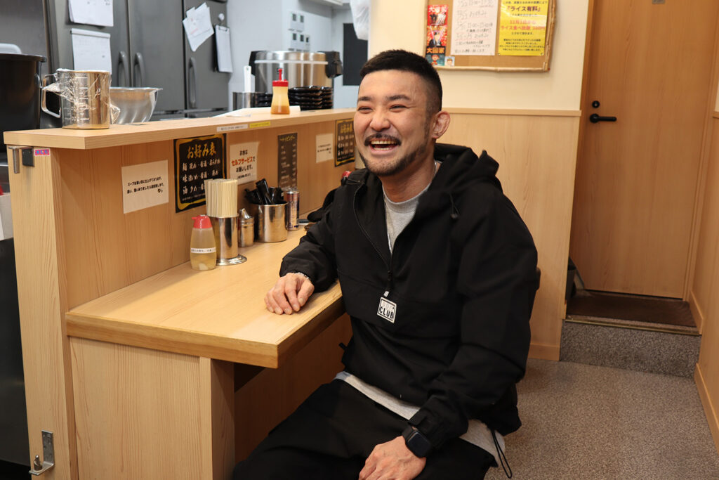 ラーメン店のカウンター席で笑顔を見せる男性の写真。男性は黒いジャケットを着てリラックスした様子で座っている。カウンターには調味料や箸が整然と並び、壁には店の案内やメニューが掲示されている。温かみのある木目調の内装が特徴で、アットホームで居心地の良さを感じさせる店内の雰囲気が伝わる。