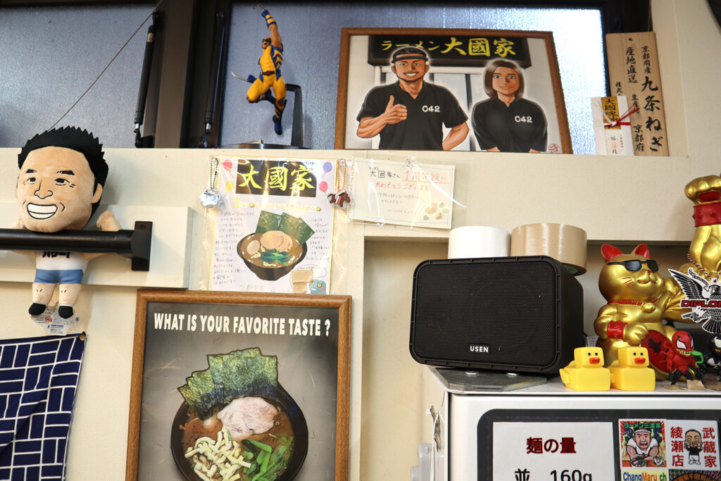 ラーメン店「大國家」の店内装飾の写真。壁には「大國家」のイラストやラーメンの写真、1周年記念ポスターが飾られている。棚の上には招き猫、黄色いアヒルのおもちゃ、スピーカーなどが置かれ、ユニークで温かみのある雰囲気が伝わる。キャラクター人形や細かい装飾品が店主の個性を反映している。