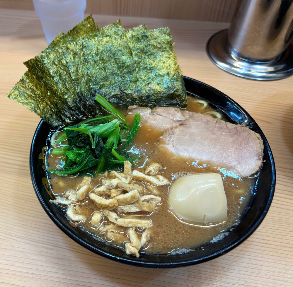 濃厚なスープの家系ラーメンの写真。黒い丼に盛られたラーメンには、大きなチャーシュー、半熟味玉、ほうれん草、刻み揚げ、そして縦に並んだ海苔がトッピングされている。スープは茶色で、しっかりとしたコクが感じられる見た目。木目調のカウンターと調味料の容器が背景に映り、ラーメン店の温かい雰囲気を伝える一枚。