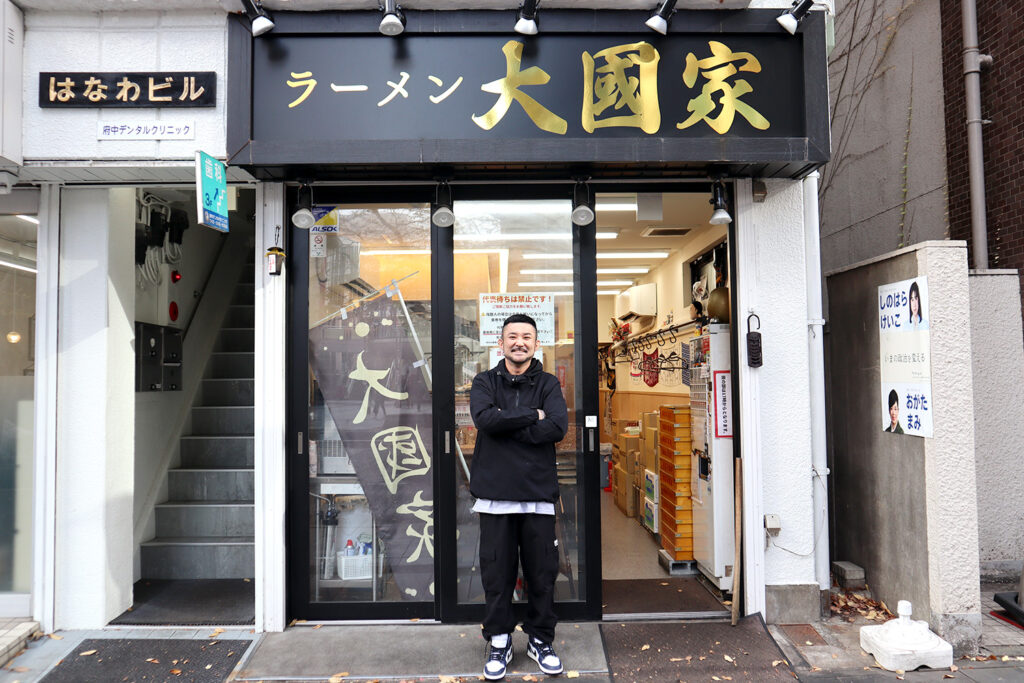 ラーメン店「大國家」の店舗前で腕を組んで立つ男性の写真。黒い看板に金色の文字で「ラーメン 大國家」と書かれており、店内がガラス越しに見える。入り口には暖簾が掛けられ、店内には調味料や道具が整然と並べられている。左側には「はなわビル」の看板と階段が写り、右側には壁に貼られた選挙ポスターが見える。親しみやすい雰囲気と店主の自信が感じられる一枚。