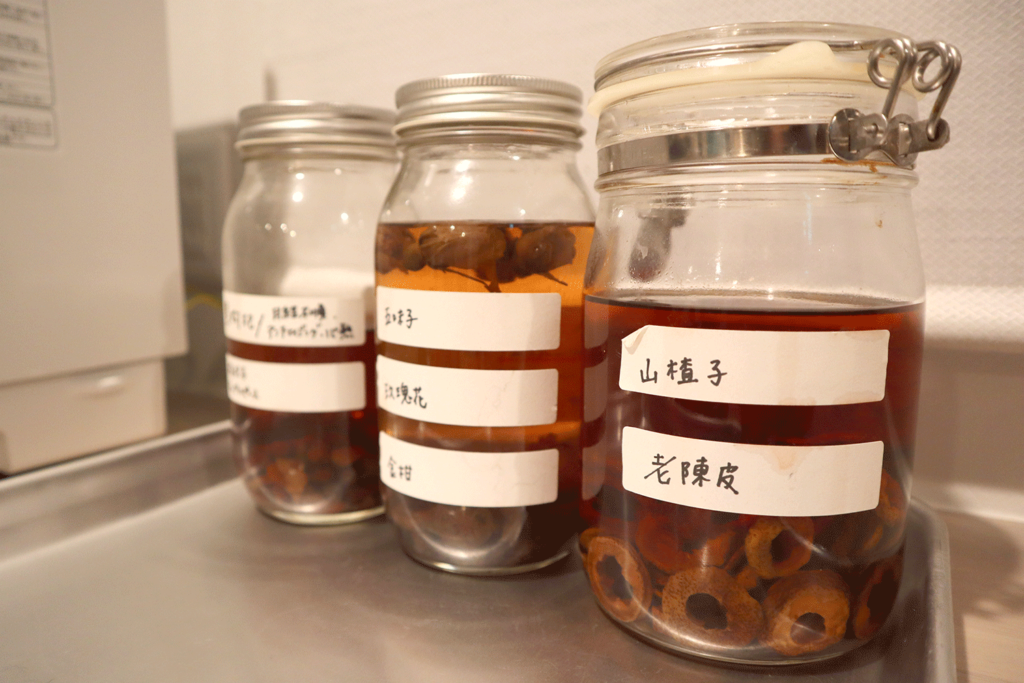 薬膳材料を漬け込んだ3本のガラス瓶がキッチンのステンレストレイの上に並んでいる。右から「山楂子(さんざし)」と「老陳皮(ちんぴ)」と書かれた瓶には輪切りの乾燥果実が漬けられ、茶色い液体に浸っている。他の瓶にも漢方素材が入っており、手書きのラベルが貼られている。丁寧に管理された薬膳素材の仕込み風景を捉えた一枚。