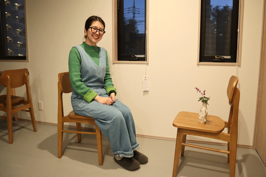 室内の窓際で、木製の椅子に座って笑顔を見せる女性。緑のニットとデニムのオーバーオールを着用し、スリッパを履いてリラックスした様子。隣の椅子には小さな花瓶に活けられた紫の花が置かれている。シンプルで温かみのあるインテリアが特徴の空間で、窓の外には夕暮れ時の景色が見える。