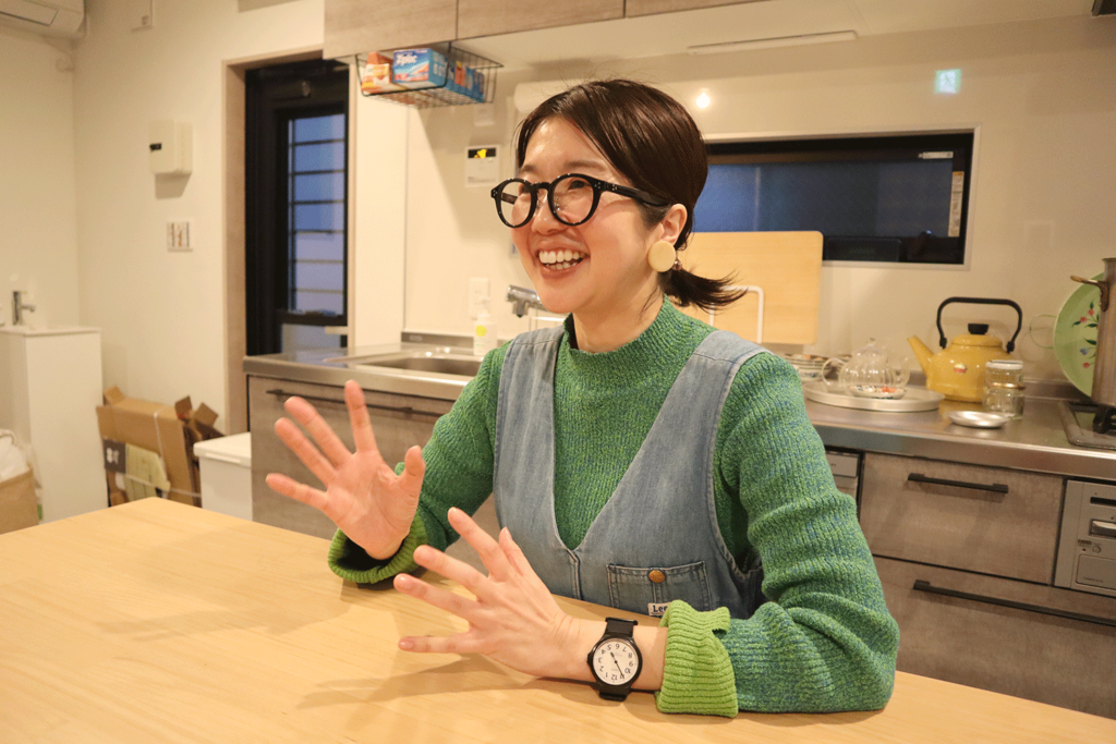 キッチンのカウンター越しに、楽しそうに話をする笑顔の女性。緑のニットにデニムのエプロンを着用し、大きめの丸い眼鏡と個性的な腕時計が印象的。背景には、黄色のやかんやガラスの急須などが置かれた整頓されたキッチンが見える。温かみのある室内で、家庭的な雰囲気が感じられる。