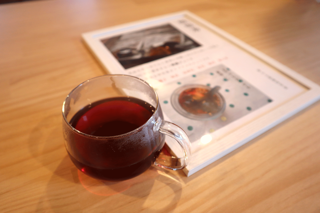 木製テーブルの上に置かれたガラスのカップに入った温かい薬膳茶。深い赤褐色の液体から湯気が立ち上り、リラックスした雰囲気を演出している。カップの奥には薬膳や料理に関する冊子が置かれており、表紙には写真と説明文が掲載されている。温かみのある空間でのひとときを感じさせる構図。
