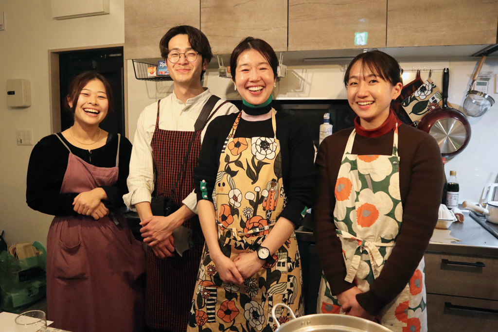 家庭的なキッチンで並んで笑顔を見せる4人の男女。全員エプロンを着用しており、料理やワークショップの準備をしている様子。中央の棚にはジップロックなどの収納アイテムが見え、後方にはフライパンや調味料が並ぶ。温かくにぎやかな雰囲気の中、チームワークの良さが感じられる集合写真。