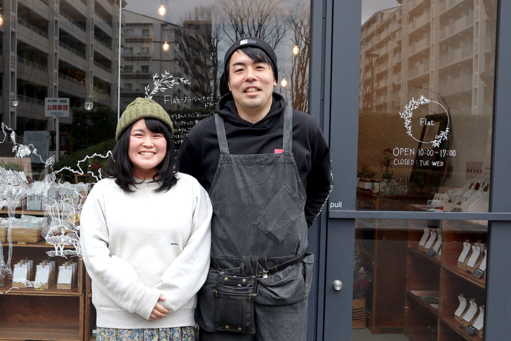 東京都府中市の「Flat」の店頭で笑顔を見せるオーナー。ガラス扉には営業時間「10:00~17:00」や定休日「火・水」が記載され、店内には商品がディスプレイされている。