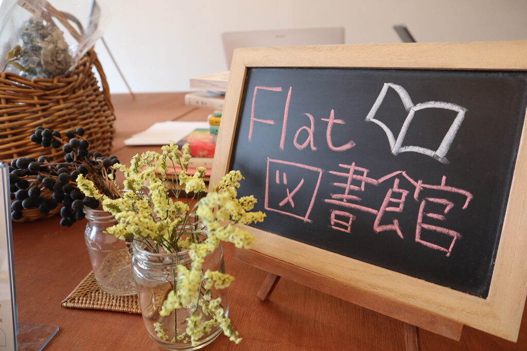 店内のテーブルに置かれた「Flat 図書館」と書かれた手書きの黒板サイン。周囲にはドライフラワーや本が並び、温かみのある落ち着いた空間が広がっている。