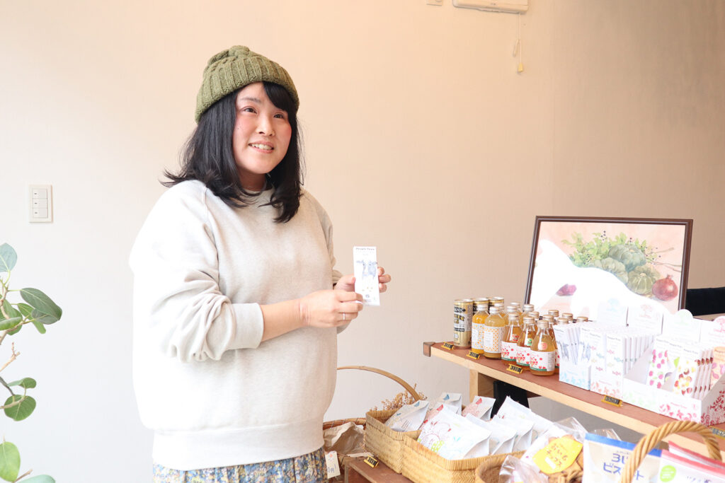 ニット帽をかぶった女性スタッフが、自然食品やドリンクが並ぶ棚の前で商品を手に持ち、笑顔で紹介している様子。