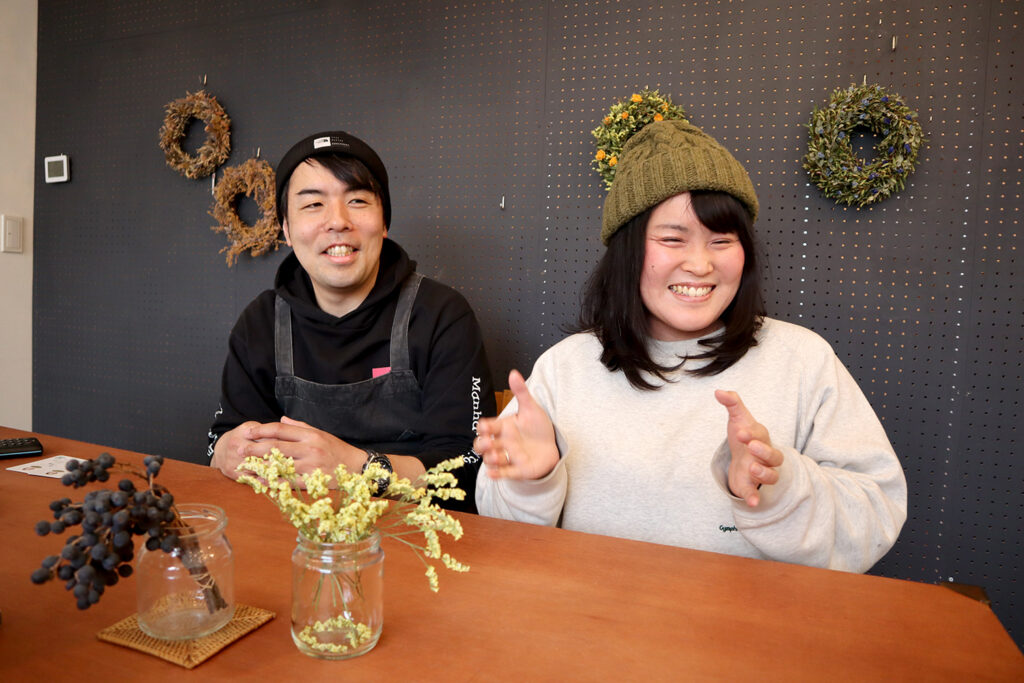 木製テーブルの向こう側で笑顔を見せる男女が座っており、背景の黒い有孔ボードにはドライフラワーのリースが飾られている、ナチュラルな雰囲気の店内風景。