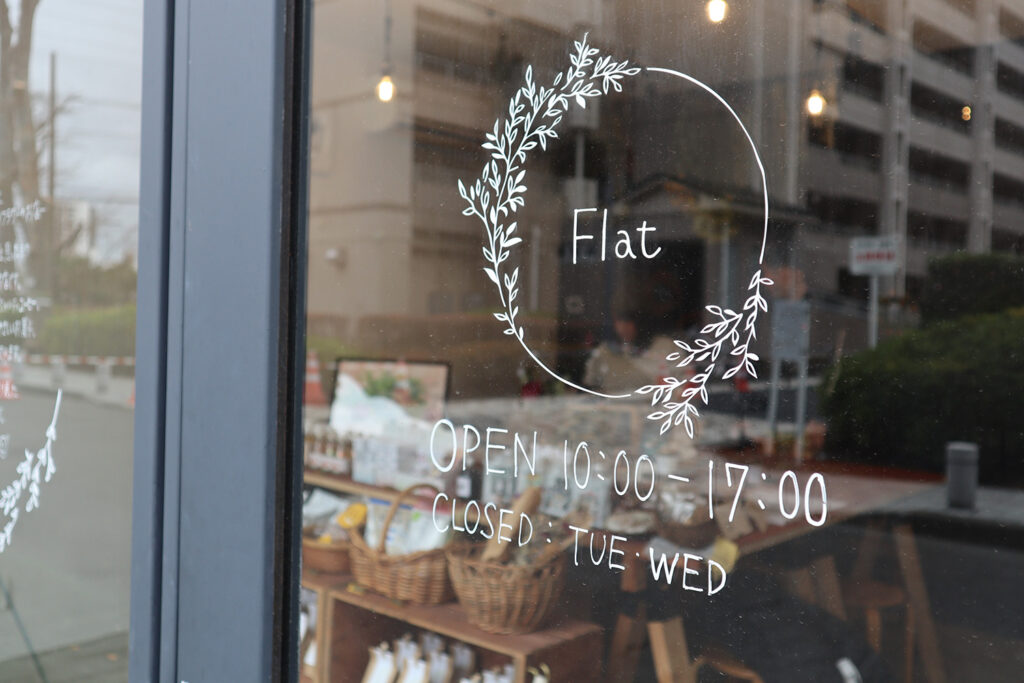 ガラス扉に描かれた「Flat」のロゴと営業時間(10:00〜17:00)、定休日(火・水)が見える。店内にはナチュラルな雑貨や商品が並び、外の街並みも反射して映っている落ち着いた雰囲気の店先の様子。