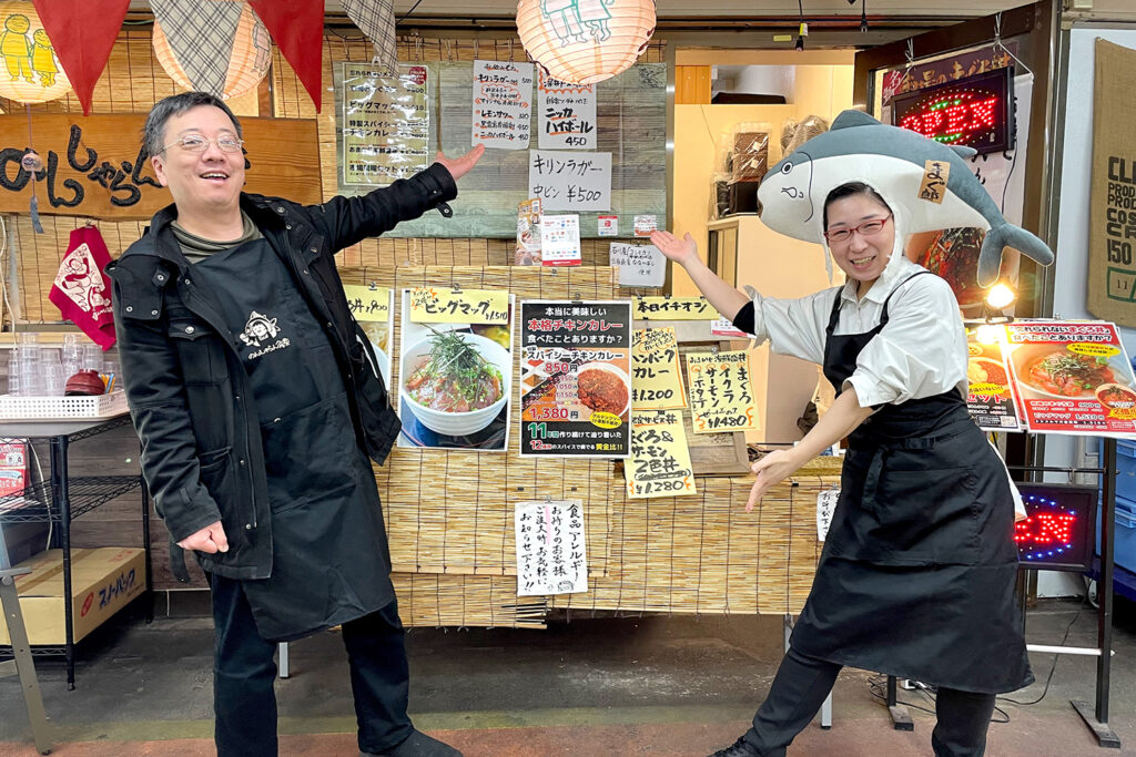 のんしゃらん食堂の前で両手を広げて笑顔を見せるスタッフ2名。おすすめメニューの看板を指差している。