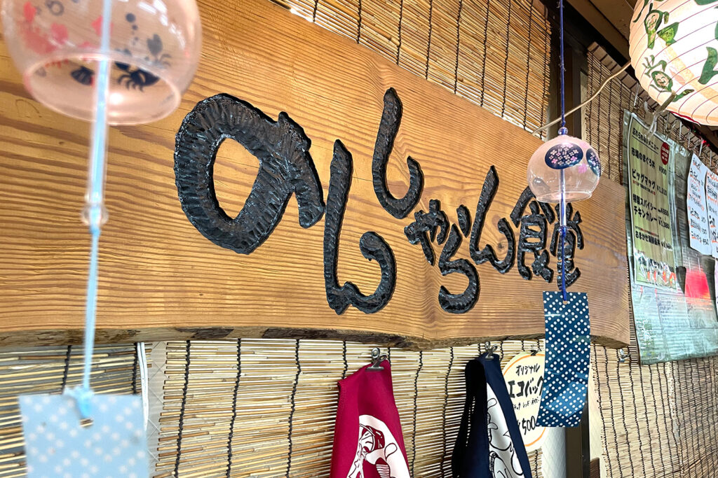 のんしゃらん食堂の木製看板のクローズアップ。風鈴と手ぬぐいが飾られ、温かみのある雰囲気を演出。