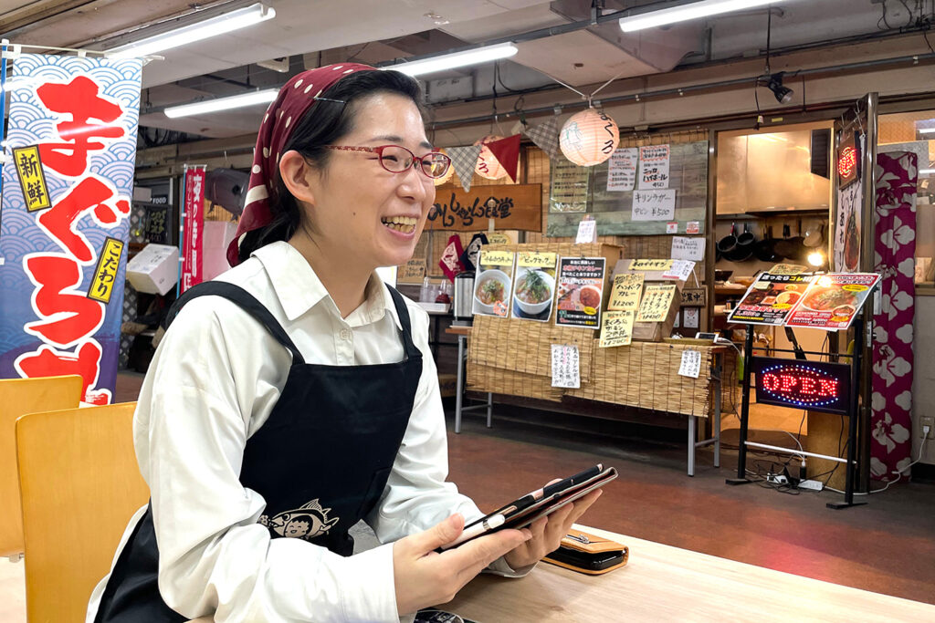 のんしゃらん食堂の店内でインタビューに応じる女性スタッフ。笑顔でタブレットを操作している様子。