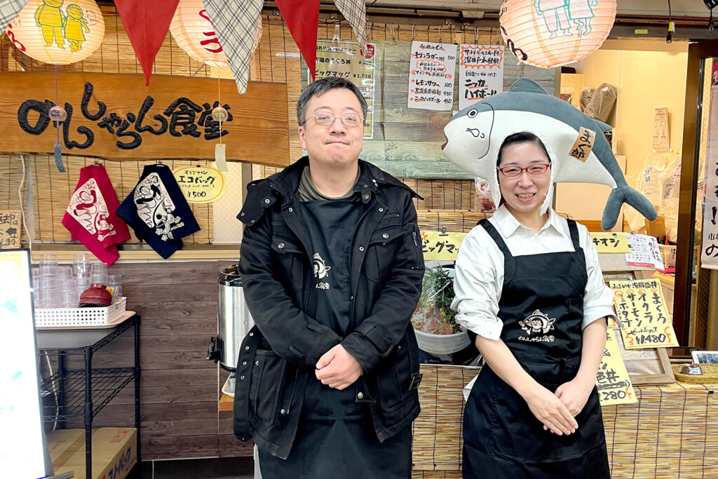 のんしゃらん食堂の店頭に立つスタッフ2名。左側の男性は黒いジャケット姿、右側の女性はマグロの被り物を着用し笑顔でポーズ。